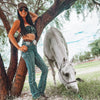 Woman in cowboy hat and jeans standing next to a white horse in a grassy area with trees.