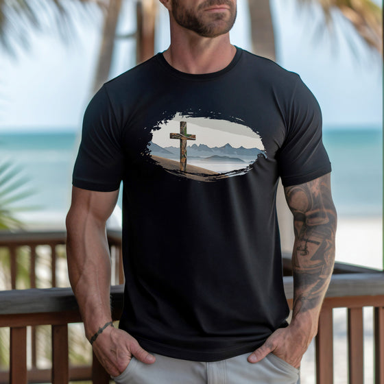 Man wearing a black t-shirt with a graphic design of a cross on a beach, standing on a wooden deck with a scenic background.