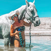 Woman in a bikini and cowboy hat standing in water with a white horse.