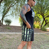 Man wearing a black tank top with white stripes and camouflage shorts with American flag design outdoors.
