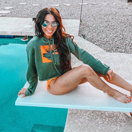 Woman at the pool on diving board wearing a River to Ridge brand cropped Elk Logo hoodie and bikini bottoms in green and orange
