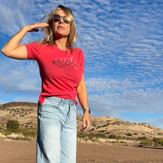 woman standing on a dirt road in new mexico wearing an elk logo t shirt and high waist jeans and crocs