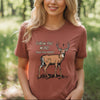 A woman with blonde hair stands in a sunlit forest clearing wearing a muted terracotta short-sleeve t-shirt with a graphic of a deer and the text 'Follow the word. Not the herd. River to Ridge Clothing