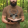 Man holding a bible in a forest wearing a brown t-shirt with a graphic design.
