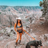 Woman in a cowboy hat with a dog on a leash, standing on a rocky trail with a canyon view.