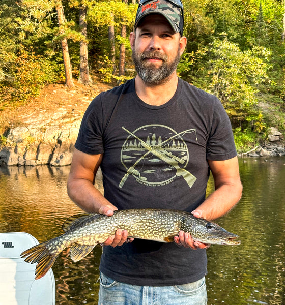 Man wearing hunting and fishing logo T shirt in black and holding a Pike fish. From River to Ridge Clothing brand