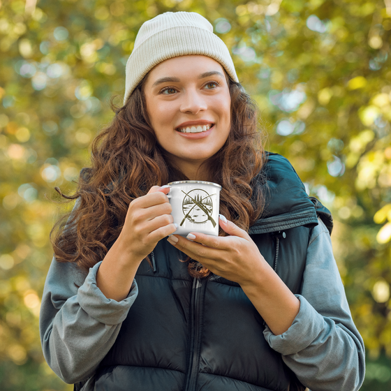 Hunting & Fishing Logo Enamel Camping Coffee Mug from River to Ridge Brand with a fishing rod and a rifle crossed over a river scene. Woman in the forest holding the mug wearing camping vest gear