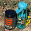 Black container with orange logo and blue water bottle with nature design on a natural background