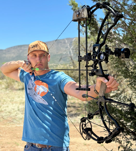 Man shooting archery and wearing a wild logo copper hat and a teal blue mountain goat logo t shirt at full draw. From River to Ridge Clothing Brand