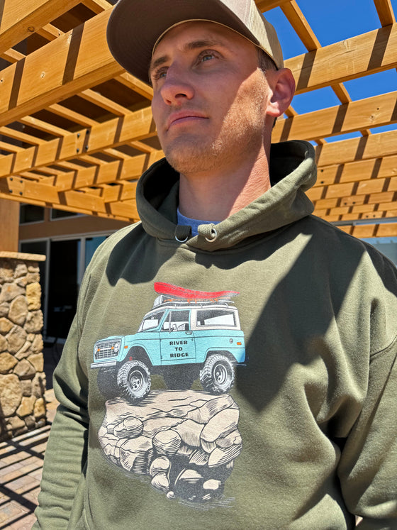 Close up of a guy wearing a River To Ridge Brand hoodie with a vintage classic Bronco on it rock crawling offroad with a red kayak tied on top of it
