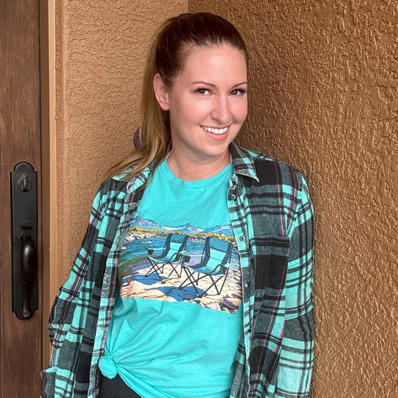 Woman wearing a teal t shirt leaning on a house and the shirt has camping chairs and a river