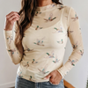 A woman wearing a beige long-sleeve mock neck top with a printed pattern of flying ducks, paired with blue jeans, in an indoor setting.