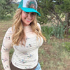 A woman outdoors wearing a teal and white trucker hat with a colorful peacock graphic, a beige long-sleeve top with a duck print, and blue jeans.