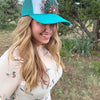 A woman outdoors wearing a turquoise and white trucker hat with a floral graphic on the front panel, paired with a light beige long-sleeve top featuring a duck print.