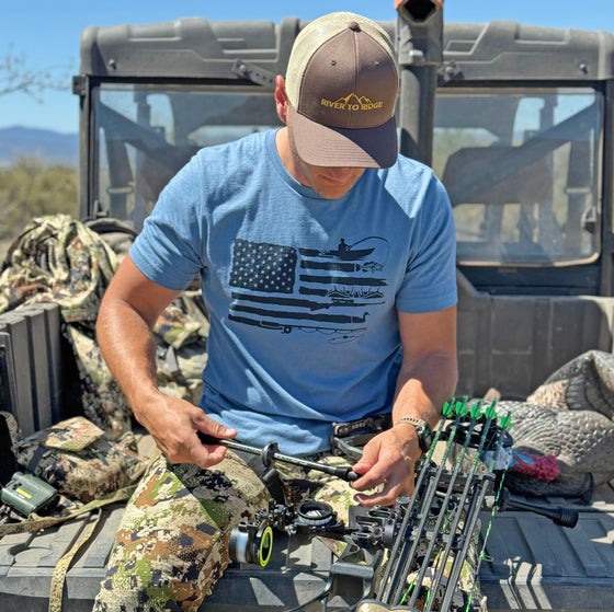 Man wearing a Sportsmans Flag T Shirt from River to Ridge clothing brand in blue and sitting on the tailgate of a 4x4 side by side working on his archery compound bow in camo pants