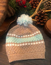 Knitted hat with striped pattern and pom-pom on a wooden surface with pine cones and branches.