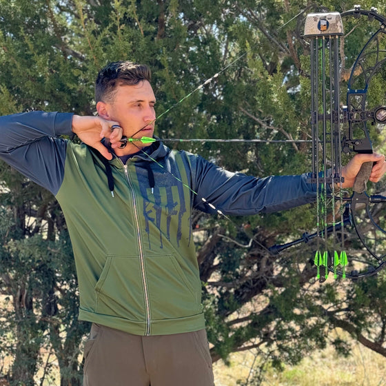 Man shooting a compound archery bow at full draw wearing a tactical logo zip up hoodie in olive and grey with rifles on it from the Brand River to Ridge Clothing