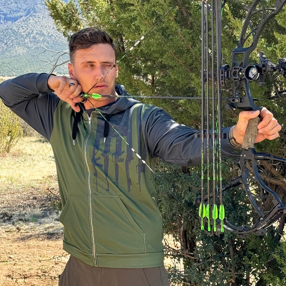 Man shooting a compound archery bow at full draw wearing a tactical logo zip up hoodie in olive and grey with rifles on it from the Brand River to Ridge Clothing