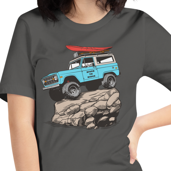 Woman wearing a River to Ridge Brand t shirt in grey with the offroad classic logo of a bronco rock crawiling with big tires and a kayak on top