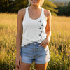 A woman standing in a sunlit grassy field wearing a white sleeveless tank top with a black bird print down the front and light blue denim shorts.